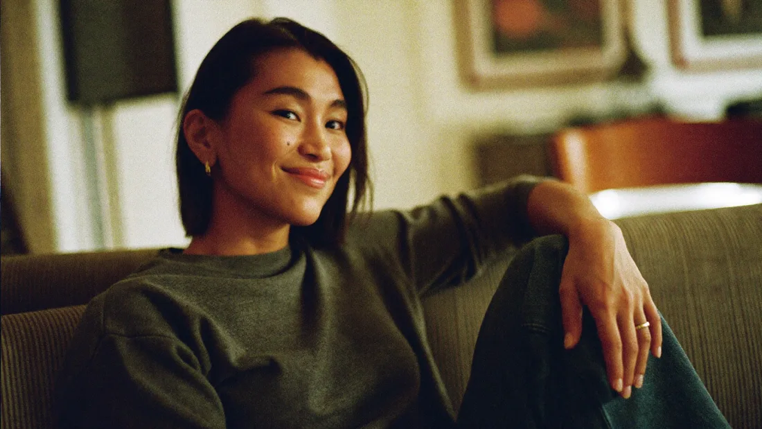 Portrait of a woman sitting back on a sofa, arm along the cushions, smiling toward the camera.