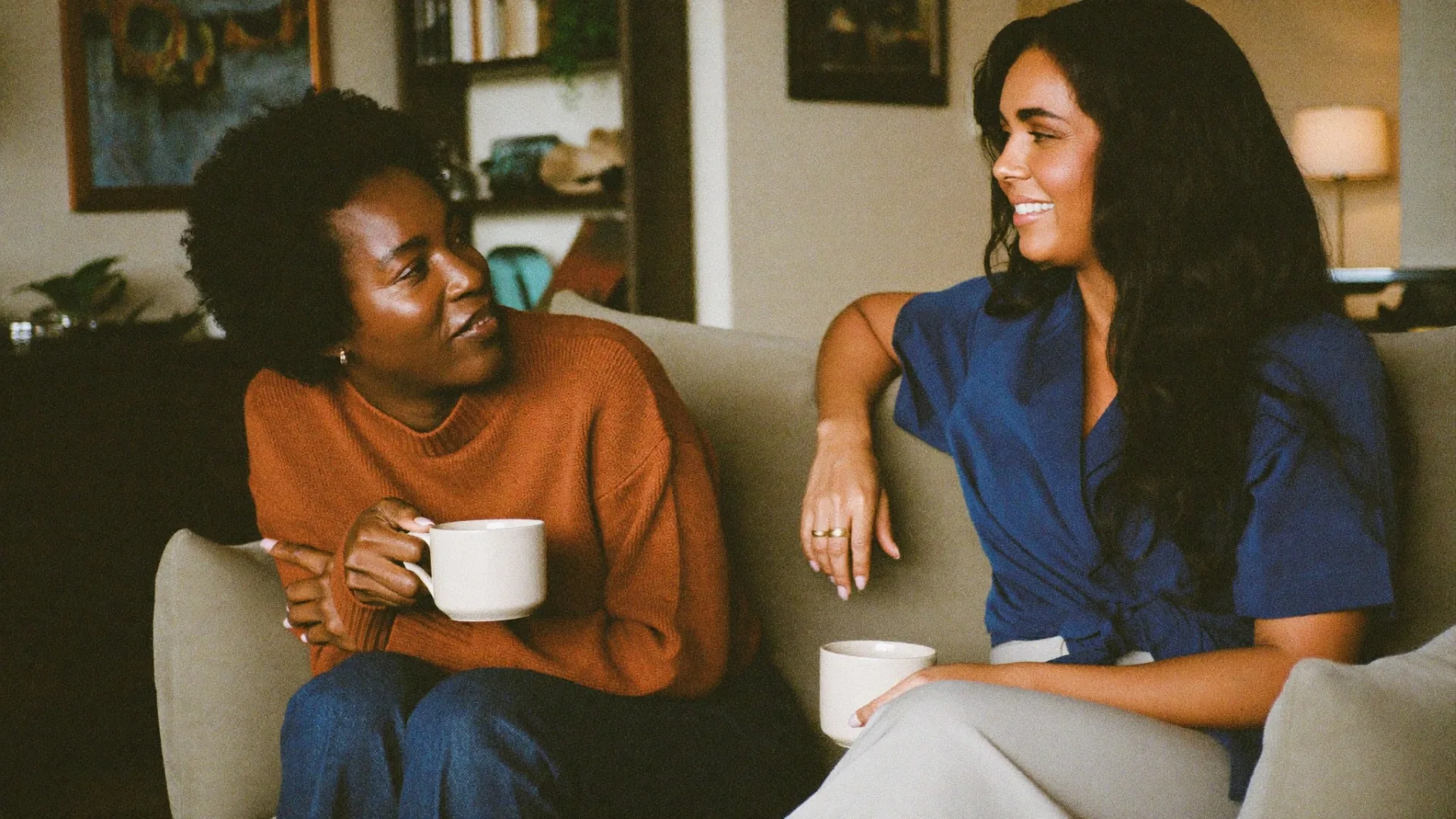 Two women sitting together with mugs, one listening while the other speaks.